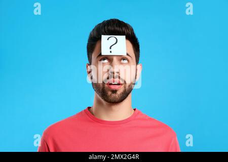 Emotional young man with question mark sticker on forehead against light blue background Stock Photo