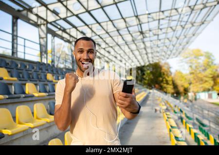 A successful man, satisfied with the result of training, looks at the camera and smiles, Hispanic holds a phone in hands uses an online sports program, an athlete holds his hand up in achievement. Stock Photo