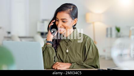 Happy business woman talking on phone call or young entrepreneur answering cellphone while sitting in front of work laptop in an office. Indian female Stock Photo