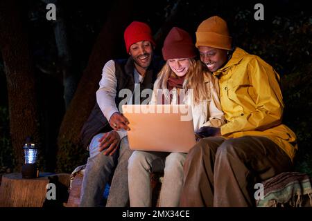 Three cheerful Mixed race Friends Using computer, sitting together at camping in the forest. Stock Photo