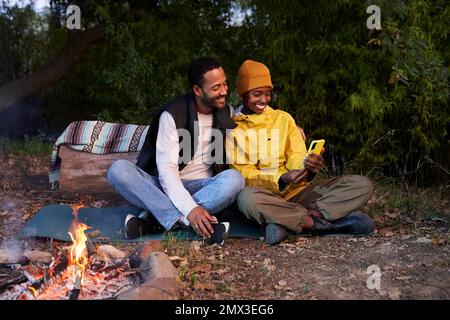 Cheerful couple sitting at camping in the mountain with campfire, using cellphone taking selfies. Stock Photo