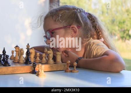 A little girl with long blonde hair and glasses is playing chess Stock Photo