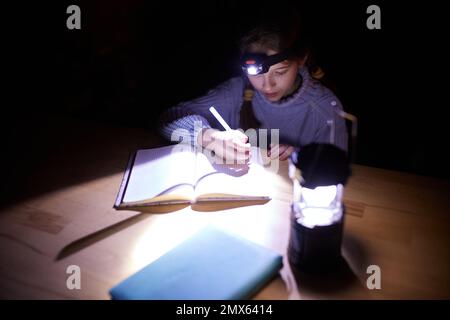 https://l450v.alamy.com/450v/2mx6414/little-school-girl-sitting-at-table-at-home-in-the-evening-and-doing-homework-without-electricity-with-battery-lamp-on-head-blackout-2mx6414.jpg