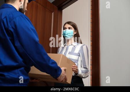 Young woman wearing medical mask receiving parcel from delivery man indoors. Prevention of virus spread Stock Photo