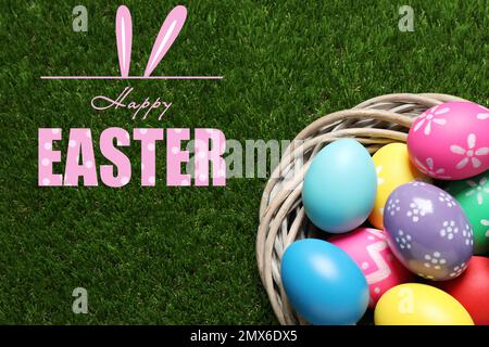 Colorful eggs in decorative nest and text Happy Easter on green grass, top view Stock Photo