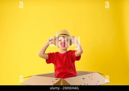 Funny little boy with clown nose in cardboard box on yellow background. April fool's day Stock Photo