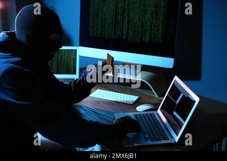 Hacker with credit card and laptop in dark room. Cyber crime Stock Photo