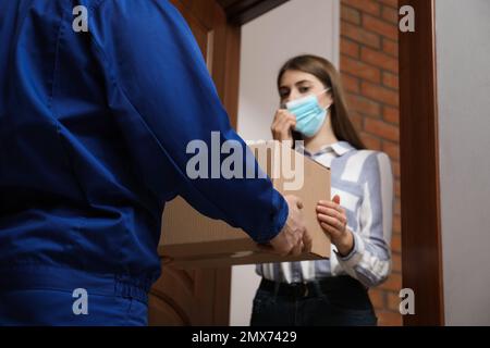 Young woman wearing medical mask receiving parcel from delivery man indoors. Prevention of virus spread Stock Photo