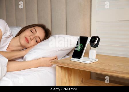 Smartphone, watch, earphones charging on wireless pad and sleeping woman in room Stock Photo