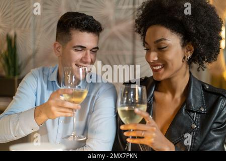 A stylish couple, him caucasian and her Afro-Italian, raises their glasses of white wine in a toast of love in a luxury restaurant setting. The scene Stock Photo