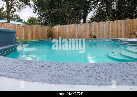 Low angle view of a large free-form gray grey accent swimming pool with turquoise blue water in a fenced-in backyard in a suburban neighborhood Stock Photo