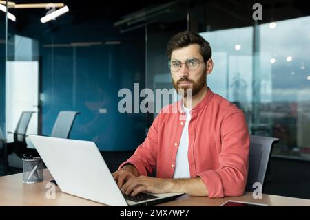 Serious and focused mature businessman in red shirt wearing glasses looking at camera, young man at work typing on laptop keyboard, programmer developer portrait at work. Stock Photo