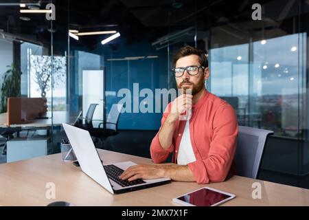 Serious and focused mature businessman in red shirt wearing glasses looking at camera, young man at work typing on laptop keyboard, programmer developer portrait at work. Stock Photo