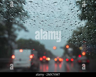 Closeup of raindrops on a windshield, and blurry road traffic with cars and red and orange lights. View from the driver's seat. Stock Photo