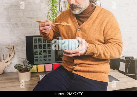 Close-up of freelancer eating oriental food, holding rice with chopsticks, resting at office desk, worker enjoying asian food at workplace during lunc Stock Photo