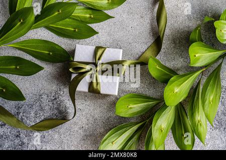 From above top view of gift box with bright green ribbon decorated with Ruscus plant leaves on concrete background Stock Photo
