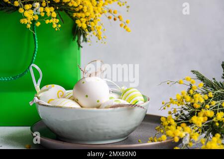 Table setting with bowl full of colored eggs with bright yellow mimosa flowers on concrete background Stock Photo