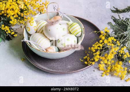 Table setting with bowl full of colored eggs with bright yellow mimosa flowers on concrete background Stock Photo