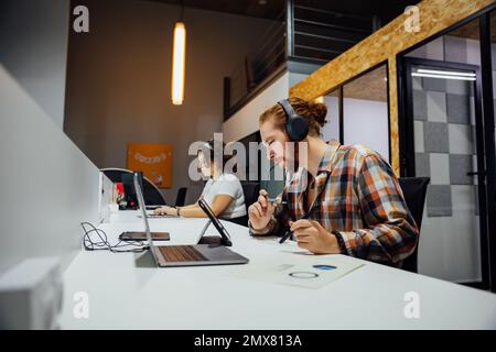 Side view of focused thoughtful bearded man in headphones with glasses in mouth looking at tablet screen while curly haired female colleague typing in Stock Photo