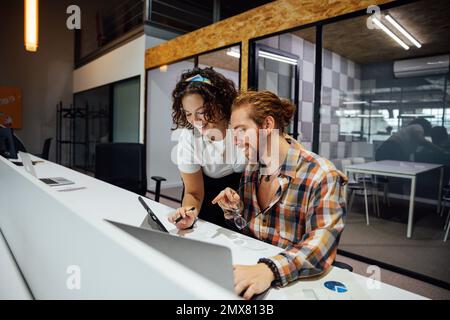 Positive bearded man in checkered shirt with glasses sitting at table and pointing at tablet screen with female colleague while collaborating on proje Stock Photo