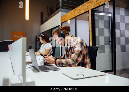 Side view of focused thoughtful bearded man in headphones with glasses in mouth looking at tablet screen while curly haired female colleague typing in Stock Photo