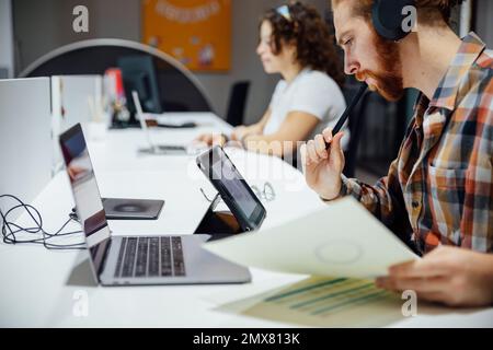 Side view of focused thoughtful bearded man in headphones with glasses in mouth looking at tablet screen while curly haired female colleague typing in Stock Photo