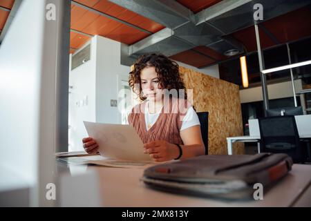 Low angle of concentrated curly haired female in casual clothes and vest sitting at table reading paper with chart while working on financial project Stock Photo