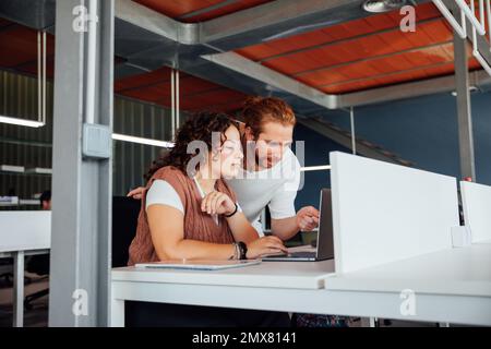 Thoughtful female sitting at table browsing on laptop with bearded male colleague collaborating on project in office Stock Photo
