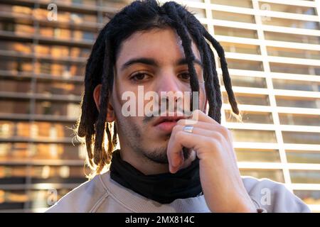 Low angle of pensive young male with dreadlocks looking at camera while touching face and standing against modern building Stock Photo