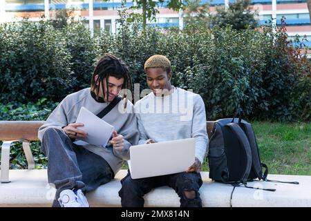 Cheerful multiethnic male friends in casual clothes sitting on bench in park and doing homework together while studying Stock Photo