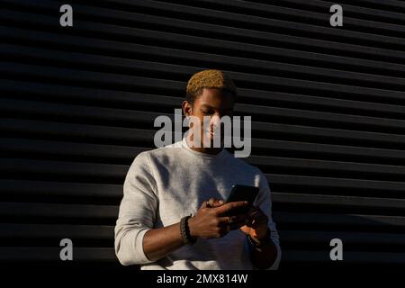Positive African American young male in casual clothes smiling and browsing smartphone while standing against black wall of modern building Stock Photo
