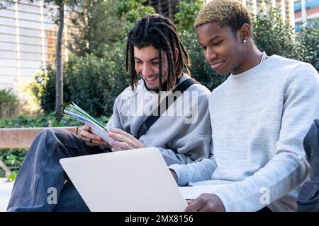 Cheerful multiethnic male friends in casual clothes sitting on bench in park and doing homework together while studying Stock Photo