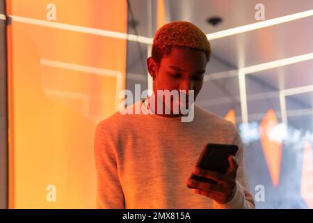 Concentrated African American young male in casual clothes messaging on smartphone while standing near wall in neon light Stock Photo