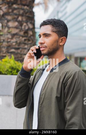 Pensive young ethnic male in casual clothing standing against modern building and speaking on mobile phone while looking away Stock Photo