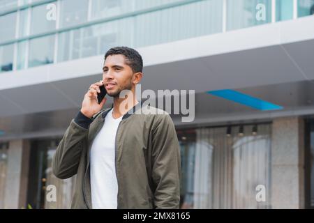 Pensive young ethnic male in casual clothing standing against modern building and speaking on mobile phone while looking away Stock Photo