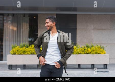 Positive young ethnic male in casual outfit smiling and looking away while standing on street Stock Photo