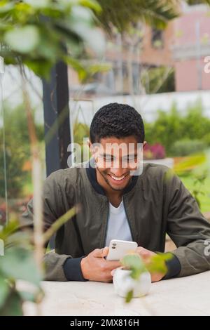 Optimistic ethnic male in casual outfit smiling and looking at screen while browsing smartphone in cafe Stock Photo