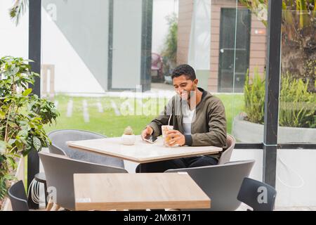 Optimistic ethnic male in casual outfit smiling and looking at screen while browsing smartphone and drinking beverage during break in earbuds in cafe Stock Photo