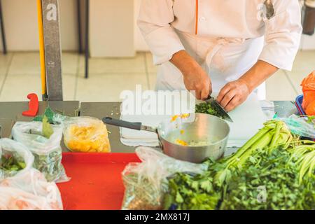 Crop anonymous chef in uniform cutting fresh green parsley with knife on cutting board in kitchen Stock Photo