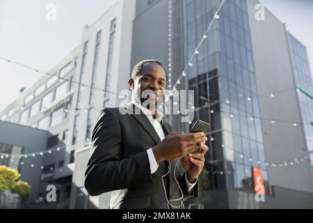 Photo from below. An African-American businessman in a suit stands outside an office center wearing headphones, holding a phone in his hand. Listens, dials, calls, He looks at the camera, smiles. Stock Photo