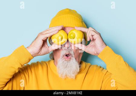 Surprised bearded elderly male in yellow sweater and hat covering eyes with lemons against blue background Stock Photo