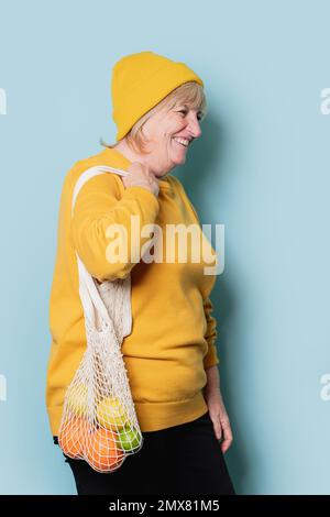Side view of positive senior female in yellow sweater and hat smiling looking away while holding net bag with fruits against blue background Stock Photo