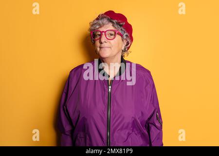 Content mature female model in glasses and beret looking at camera while standing against yellow background in colorful clothes Stock Photo
