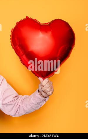 Unrecognizable senior man in pink clothes with red heart shaped balloon on yellow background Stock Photo
