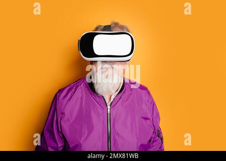 Portrait of bearded elderly male wearing VR goggles and purple jacket standing against orange background while exploring cyberspace in studio Stock Photo