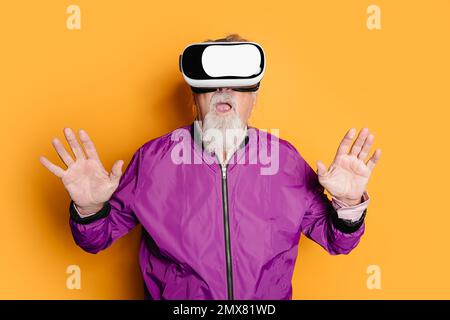 Amazed senior male in bright clothes experiencing virtual reality with raised hands while standing against yellow background in VR goggles Stock Photo