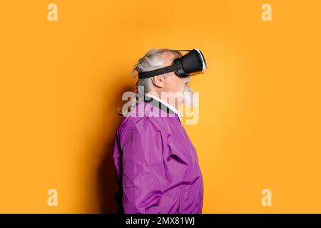 Portrait of bearded elderly male wearing VR goggles and purple jacket standing against orange background while exploring cyberspace in studio Stock Photo