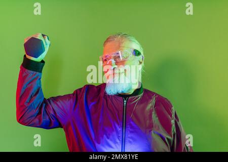 Serious bearded senior male hipster in casual clothes and glasses standing against green background while lifting dumbbell Stock Photo