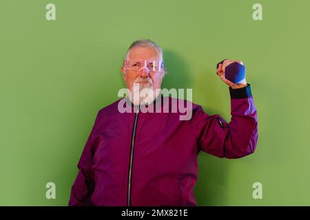 Serious bearded senior male hipster in casual clothes and glasses standing against green background while lifting dumbbell Stock Photo