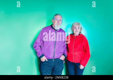 Confident senior couple in bright clothes and glasses standing against green wall and looking away thoughtfully Stock Photo